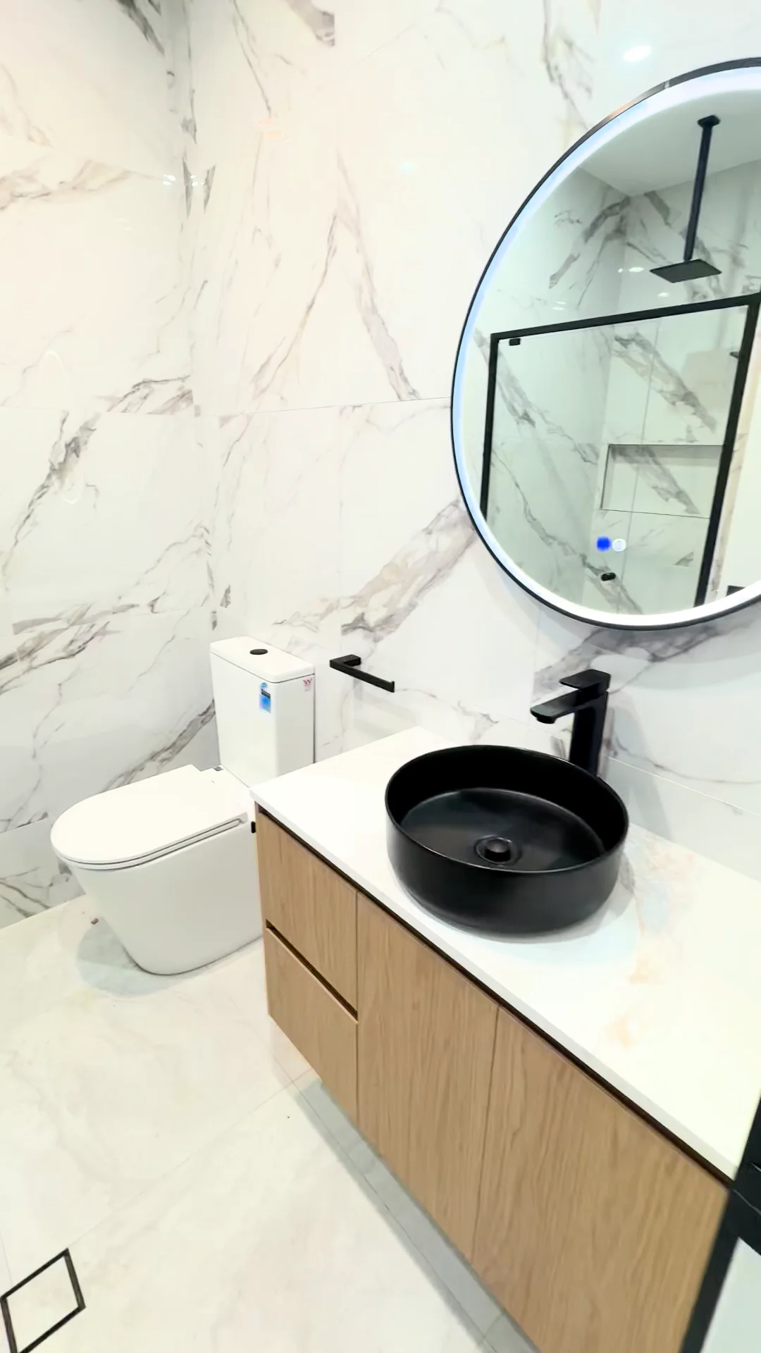 Austral bathroom with an illuminated round mirror, black basin and tapware, timber vanity and pale veined tiles
